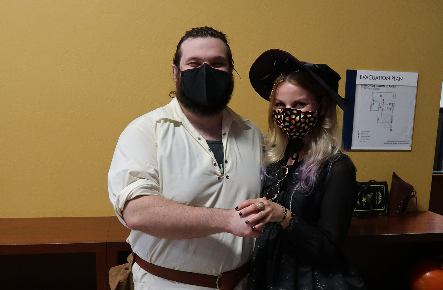 Student wearing a beige button-up and black mask smiling and holding hands with student wearing a black hat and candy corn mask.