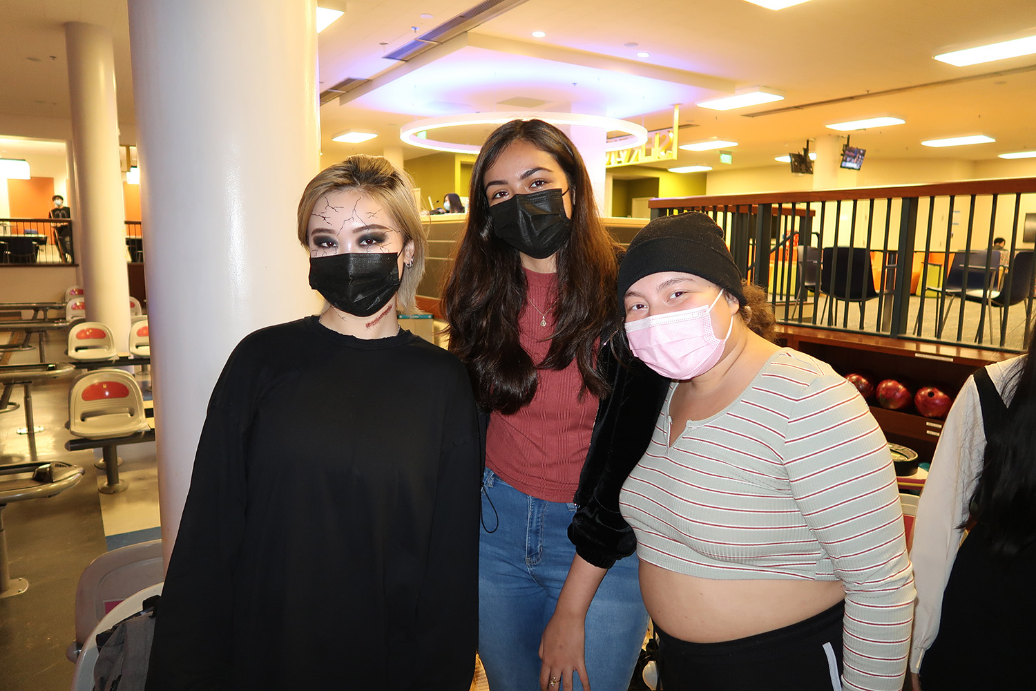 Three students with masks smiling. Leftmost student has blood on their neck, black eyeshadow, and black cracks on their forehead.