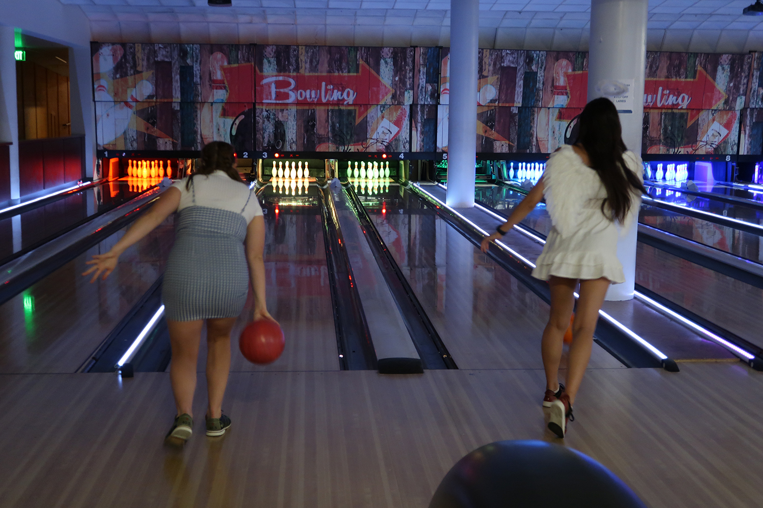 Two students throwing bowling balls in the UCD Game Room. Left student is wearing a blue plaid dress and white shirt. Right student is wearing a white dress with ruffled skirt.